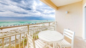 Varanda com mesa e cadeiras brancas com vista para uma praia, com pessoas e guarda-sóis.