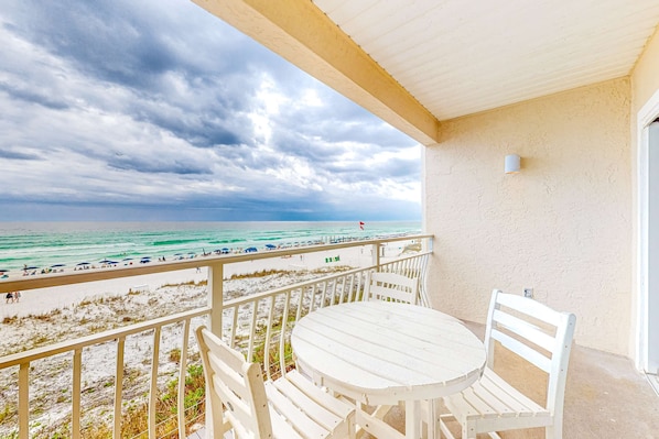 Ein Balkon mit weißem Tisch und Stühlen mit Blick auf einen Strand, auf dem sich Menschen und Sonnenschirme befinden.