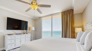Ein Hotelzimmer mit einem großen Bett, einem Deckenventilator, einem Flachbildfernseher, einer weißen Kommode, einem Balkon mit Meerblick und einem großen Fenster mit Vorhängen.