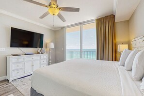 A hotel room with a large bed, a ceiling fan, a flat-screen TV, a white dresser, a balcony with a view of the ocean, and a large window with curtains. - Eighth-floor beachfront condo w/ hot tubs, pools, seasonal beach service & more (Panama City Beach)