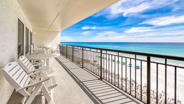 A balcony with white chairs and a table, overlooking a beach with umbrellas and a clear blue sky. - 6th-floor, updated condo with beach service - near dining (Fort Walton Beach)