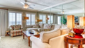 A spacious living room with a large sofa, a smaller armchair, a coffee table, and a TV. There are two ceiling fans, a large mirror, and a window with blinds. - 6th-floor, updated condo with beach service - near dining (Fort Walton Beach)