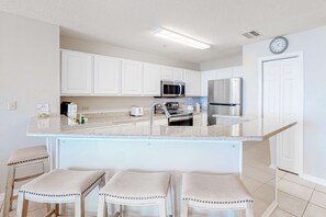 Una cocina con armarios blancos, electrodomésticos de acero inoxidable, encimera de mármol y tres taburetes de bar.