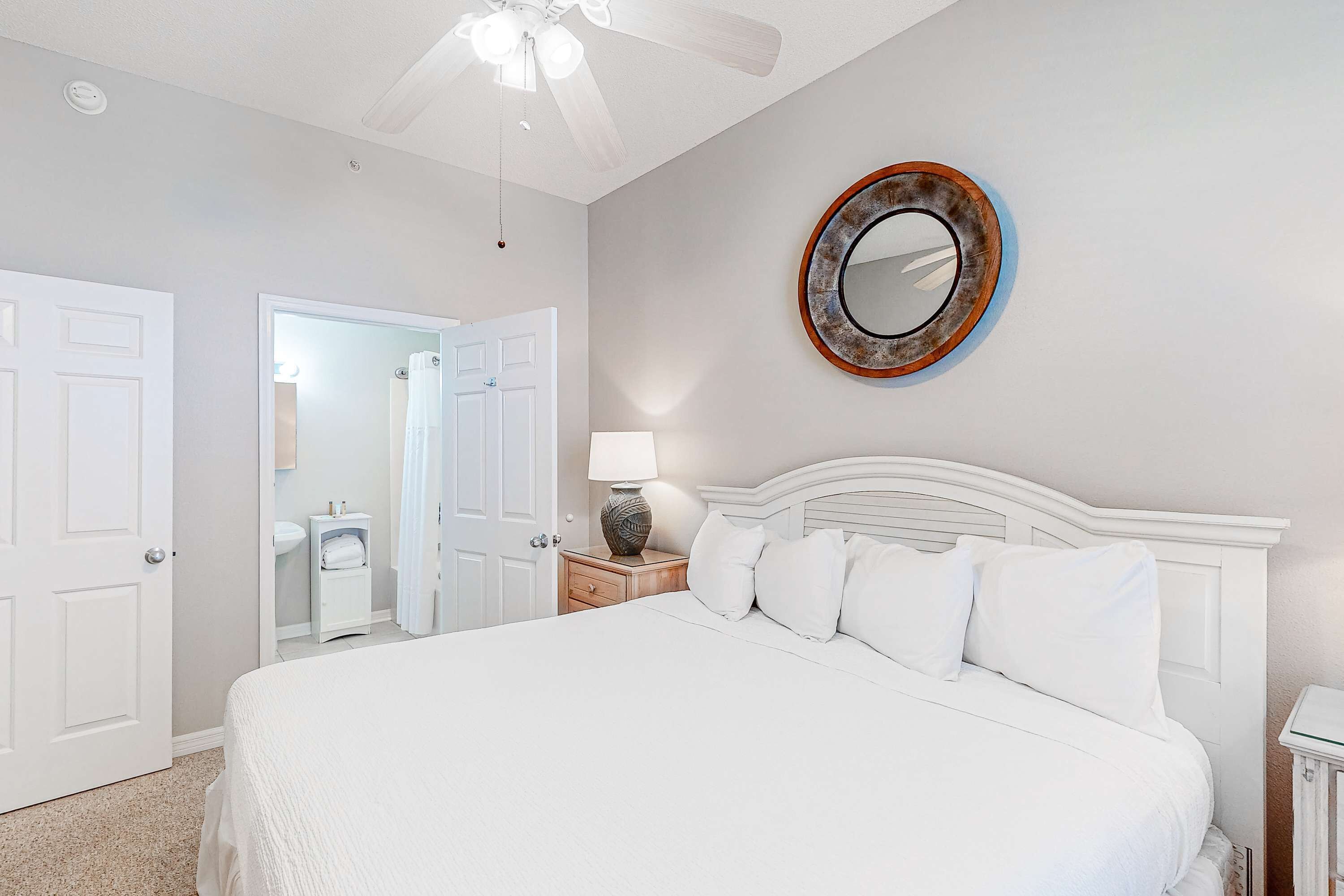 Une chambre à coucher avec un grand lit, un ventilateur de plafond, un miroir rond et une table de chevet avec une lampe.
