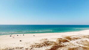 Ein Strand mit weißem Sand und klarem, blauem Wasser. Mehrere Liegestühle und Sonnenschirme sind vorhanden.