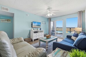 Ein Wohnzimmer mit einem weißen Sofa, einem blauen Sofa, einem Couchtisch und einem Deckenventilator. An der Wand hängt ein Gemälde und das Fenster bietet einen Blick ins Freie.