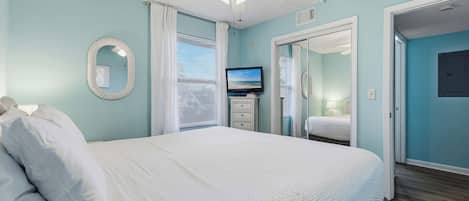 Une chambre à coucher avec un lit, un ventilateur de plafond, un téléviseur et une salle de bain visible à travers une porte ouverte.