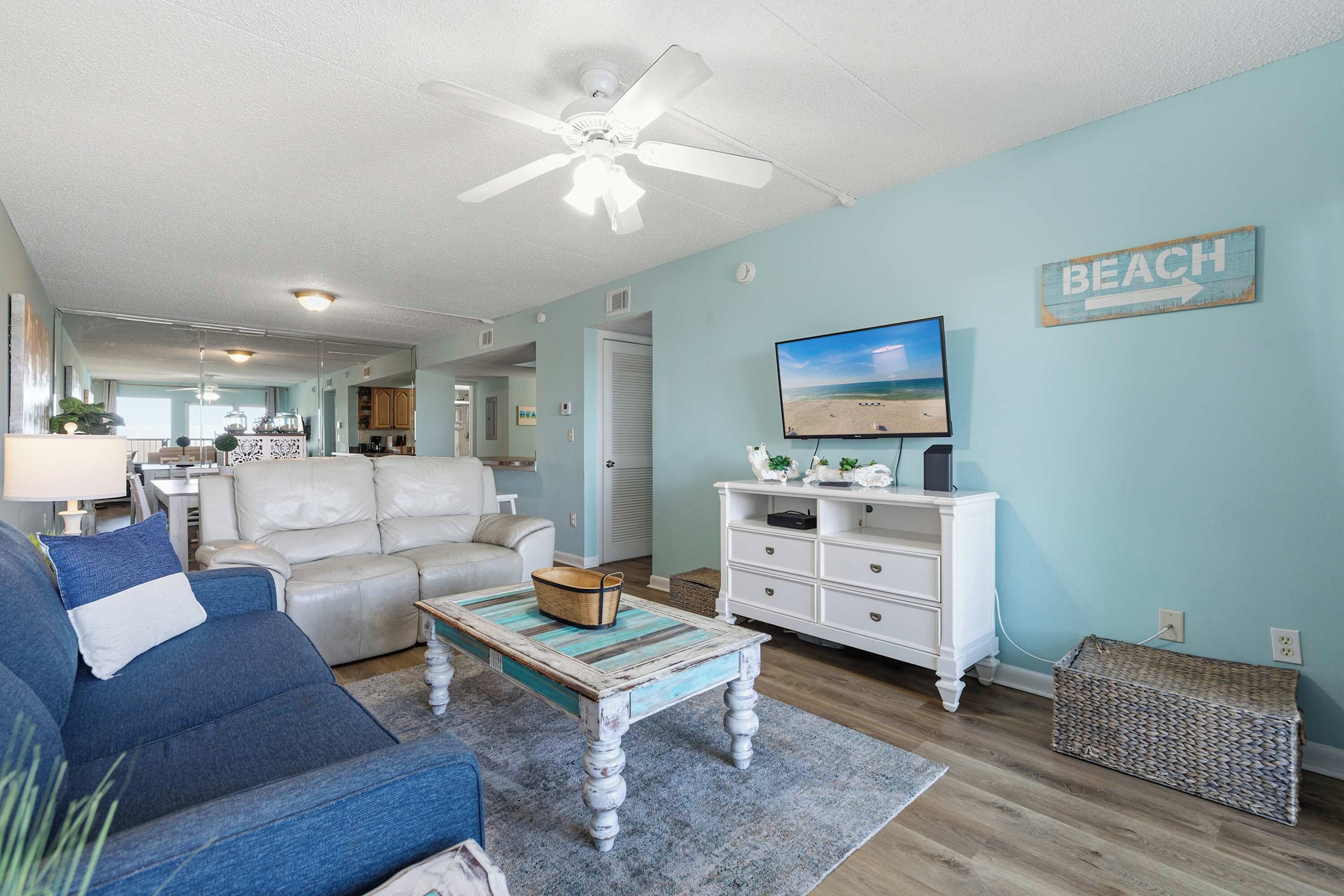A living room with a ceiling fan, a television on a white dresser, and a 'BEACH' sign on the wall.