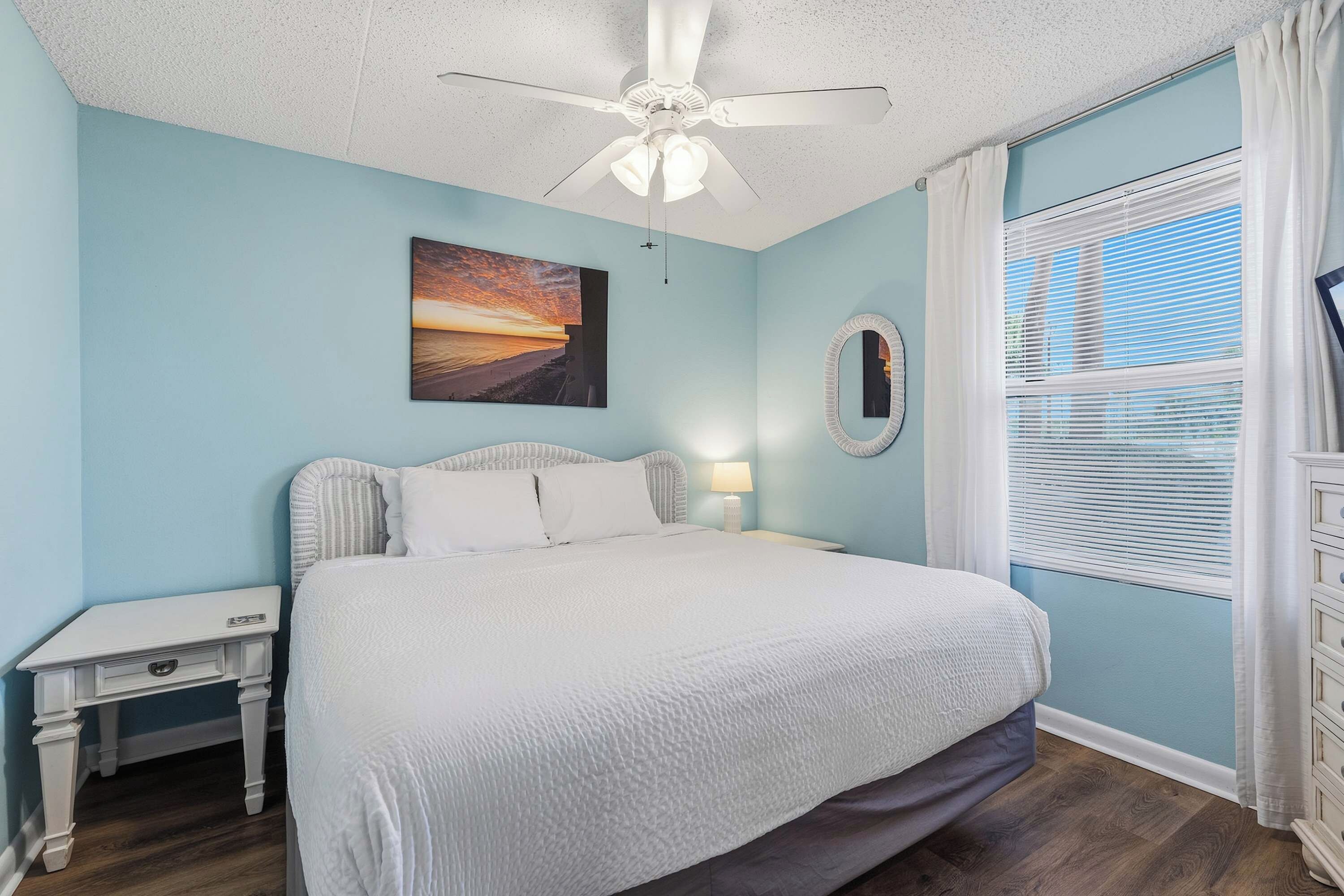 Une chambre à coucher comprenant un lit, une table de chevet, un ventilateur de plafond, un tableau, un miroir et une fenêtre avec des stores.
