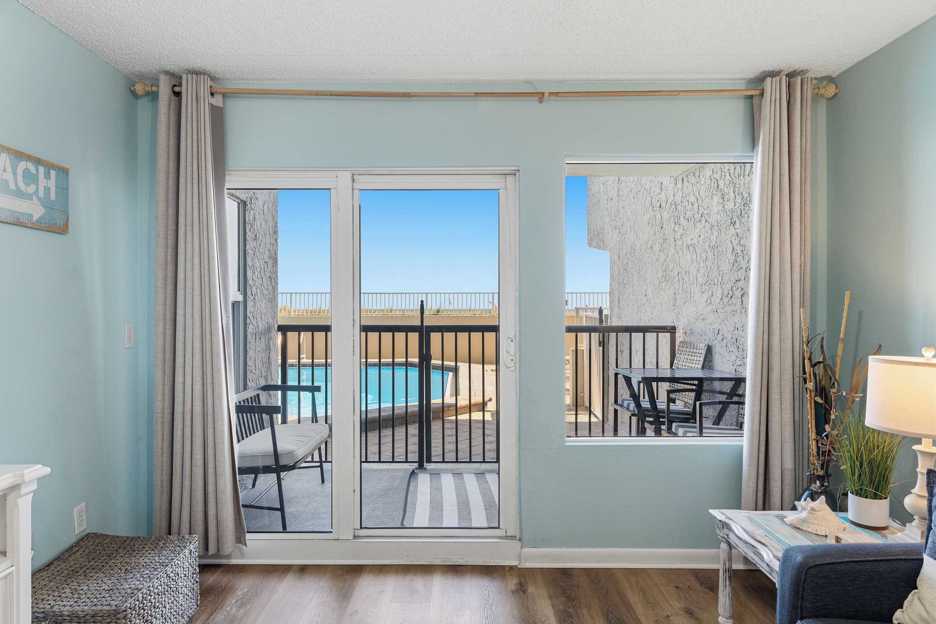 A room with a view of a pool and a beach, a table with chairs, and a lamp on a side table.