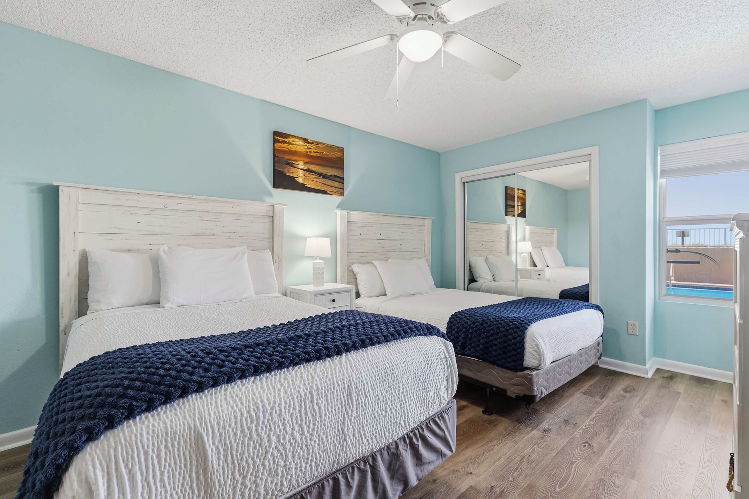 A hotel room with two beds, a ceiling fan, a large mirror, and a view of the outdoors.