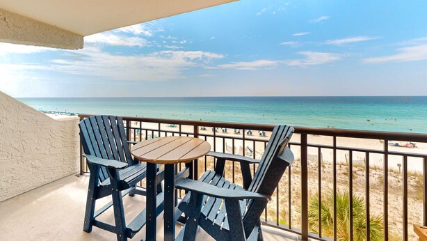 A balcony with two Adirondack chairs and a small table, overlooking a beach with a clear view of the ocean and distant shoreline. - Cozy Gulf-front condo with beach service, pool, tennis & fitness room (Fort Walton Beach)