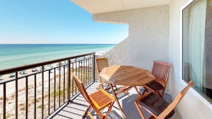 A balcony with a wooden table and chairs, overlooking a beach with umbrellas and people. - Updated, bright condo with beach setup - minutes from entertainment (Fort Walton Beach)