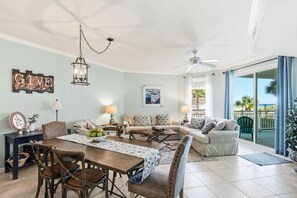 A spacious living room with a dining area, a ceiling fan, a large window with a view of the beach, and a 'GIVE thanks' sign on the wall. - Charming two-bedroom with bunks, balcony overlooking pool & pickleball (Santa Rosa Beach)
