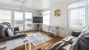 Un salon spacieux avec un grand canapé, une table basse et un téléviseur. De grandes fenêtres sont équipées de stores, et on y trouve également un radiateur et une horloge murale.