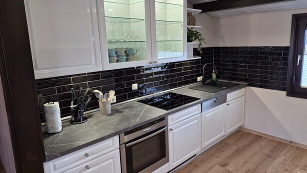 A modern kitchen with white cabinets, black tiled backsplash, stainless steel appliances, and a glass-fronted upper cabinet. - Freistehender Ferienbungalow im Fachwerkstil (Burhave)