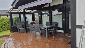 A covered patio with a table and chairs, a grill, and a view of the garden. - Freistehender Ferienbungalow im Fachwerkstil (Burhave)
