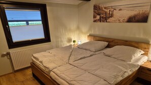 A bedroom with a bed, a window, a radiator, and a painting on the wall. - Freistehender Ferienbungalow im Fachwerkstil (Burhave)