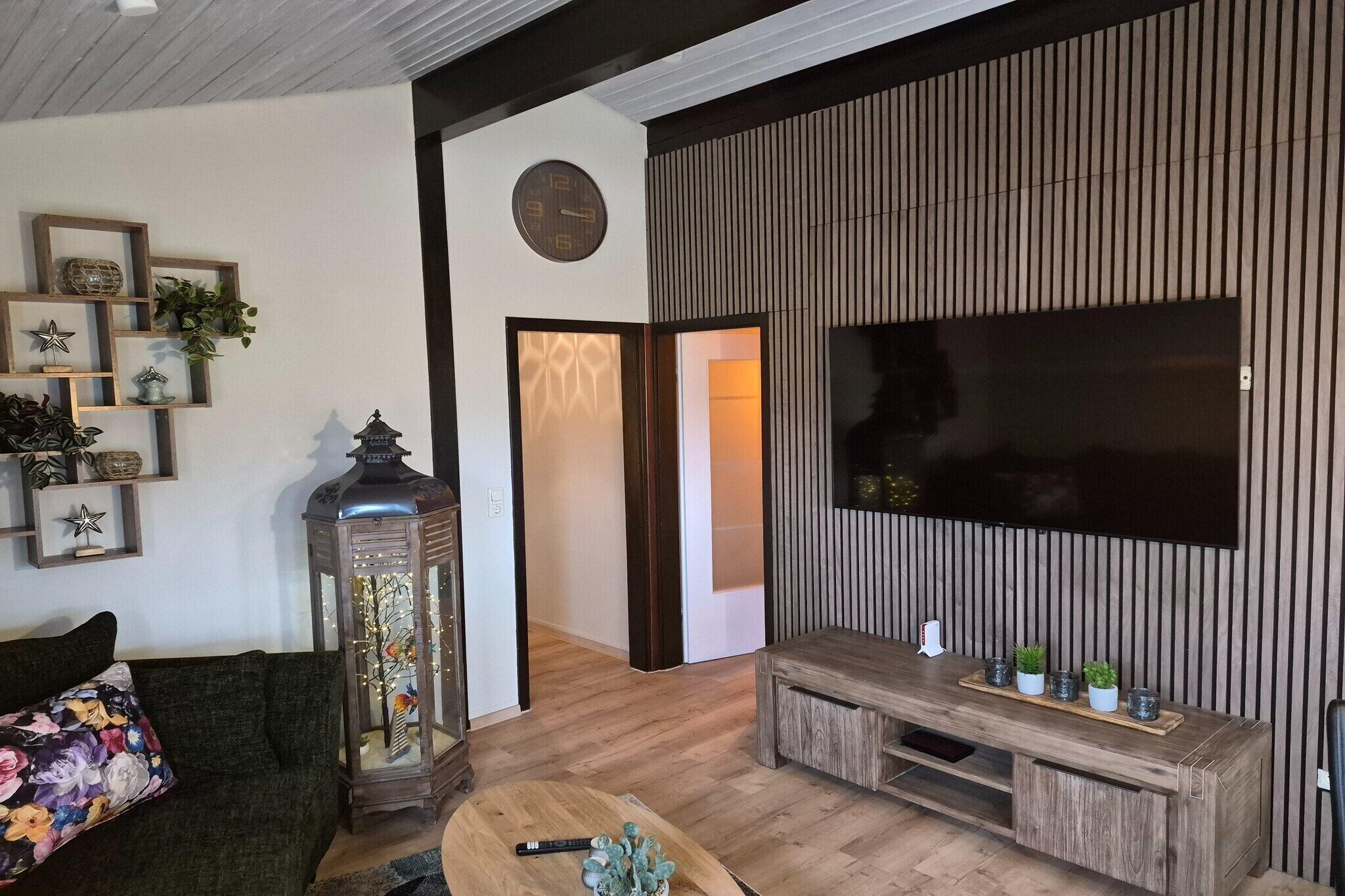 Una sala de estar moderna con piso de madera, un televisor de pantalla plana grande montado en una pared de paneles de madera, un sofá verde oscuro con un cojín floral, una mesa de centro de madera y un farol decorativo.