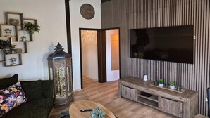 A modern living room with a wooden floor, a large flat-screen TV mounted on a wooden panel wall, a dark green sofa with a floral cushion, a wooden coffee table, and a decorative lantern. - Freistehender Ferienbungalow im Fachwerkstil (Burhave)