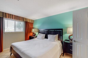 A hotel room with a large bed, two bedside tables with lamps, a window with curtains, and a teal-colored wall. - Oceanfront Condo with AC, WiFi, Shared Pool, & Seasonal Beach Service Rentals (Destin)