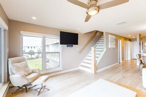 A spacious living room with a large window, a ceiling fan, a staircase, and a flat-screen TV mounted on the wall. - Ramsgate getaway w/ panoramic view, beach access & shared pool (Panama City Beach)