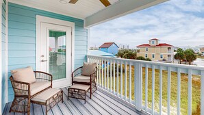 Eine Veranda mit Korbstühlen und Deckenventilator, mit Blick auf ein Wohngebiet mit Häusern und einem Strand.