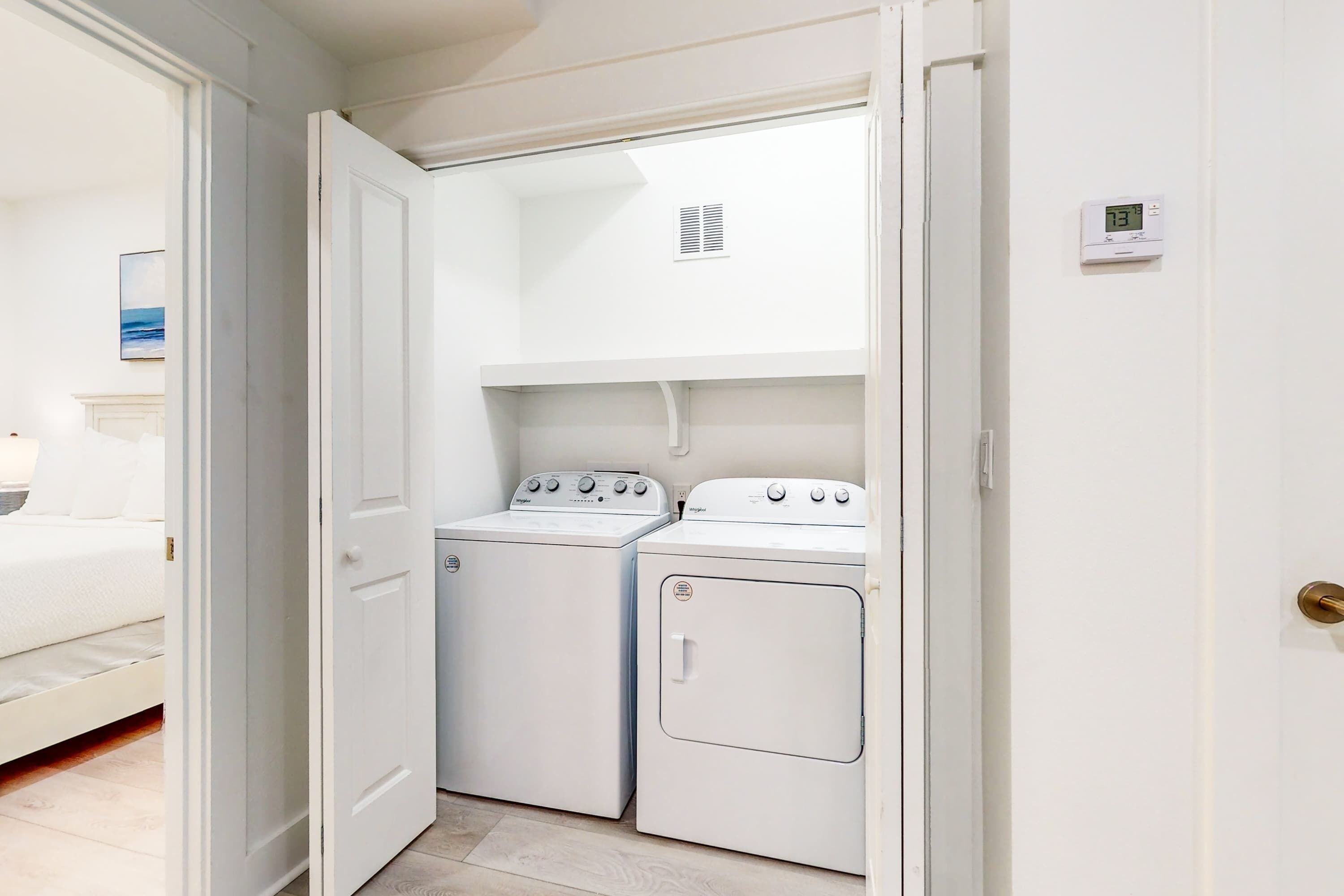 A laundry room with two white appliances, a bed in the adjacent room, and a thermostat on the wall.