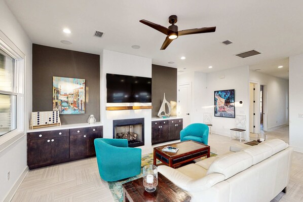A modern living room with a fireplace, teal chairs, a white sofa, and a ceiling fan. - Home near beach and 30A with balcony, yard, patio, grill, Washer & Dryer (Santa Rosa Beach)