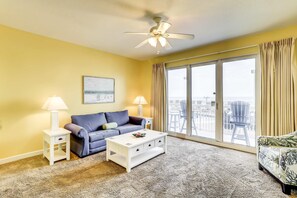 Ein Wohnzimmer mit einer Couch, einem Couchtisch und einem Deckenventilator. Es gibt ein großes Fenster mit Vorhängen und Blick auf die Stadt.