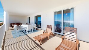 Un balcone con tavolo e sedie in legno, una poltrona prendisole in legno e vista sull'oceano attraverso ampie finestre.