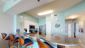 A modern kitchen with a bar area, featuring a granite countertop, stainless steel appliances, and bar stools with wooden accents. - Updated oceanfront condo with pools, hot tub, W/D, AC, & views (Panama City Beach)