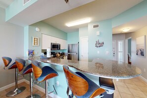 A modern kitchen with a bar area, featuring a granite countertop, stainless steel appliances, and bar stools with wooden accents. - Updated oceanfront condo with pools, hot tub, W/D, AC, & views (Panama City Beach)
