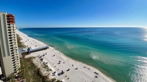 A beach with a hotel building on the left, clear blue water, and a sandy shore. - Updated oceanfront condo with pools, hot tub, W/D, AC, & views (Panama City Beach)