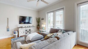 A living room with a gray sectional sofa, a ceiling fan, a flat-screen TV, a white cabinet, and a window with blinds. - Ideal Coastal Retreat w/ Shared Pool, Private Balcony, & Beach Access Nearby (Panama City Beach)