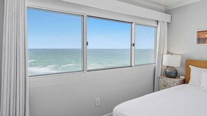 A hotel room with a large window overlooking the ocean, a bed with white bedding, a nightstand with a lamp, and a wooden headboard. - Gorgeous beachfront condo, shared pool and beach access (Panama City Beach)