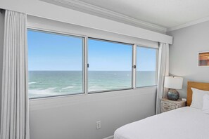 A hotel room with a large window overlooking the ocean, a bed with white bedding, a nightstand with a lamp, and a wooden headboard. - Gorgeous beachfront condo, shared pool and beach access (Panama City Beach)