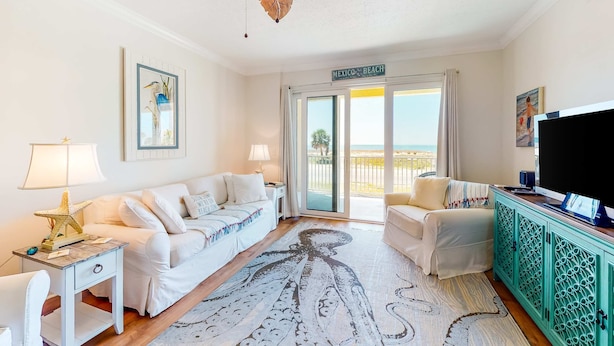 Een goed verlichte kamer met een plafondventilator, een bed, een bank, een televisie en uitzicht op het strand.