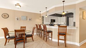 A dining area with a round glass table and wooden chairs, a kitchen with white cabinets and a black microwave, and a clock on the wall.