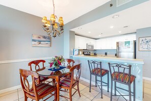 A dining area with a wooden table and chairs, a chandelier, and a kitchen in the background. - Cozy, family-friendly beach condo w a furnished balcony, shared pool, tennis (Destin)