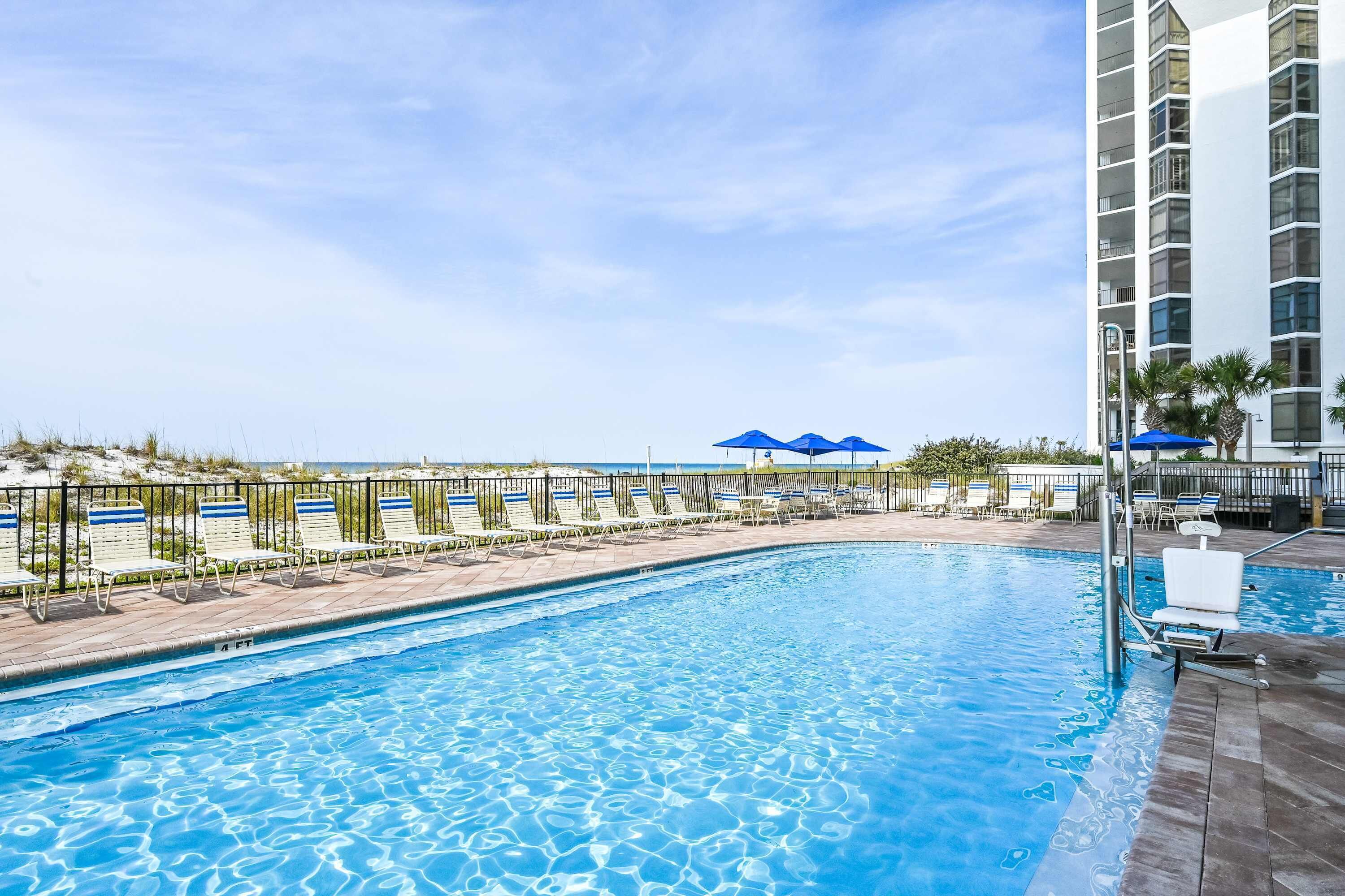 A swimming pool with lounge chairs and umbrellas, surrounded by a fence and a building.