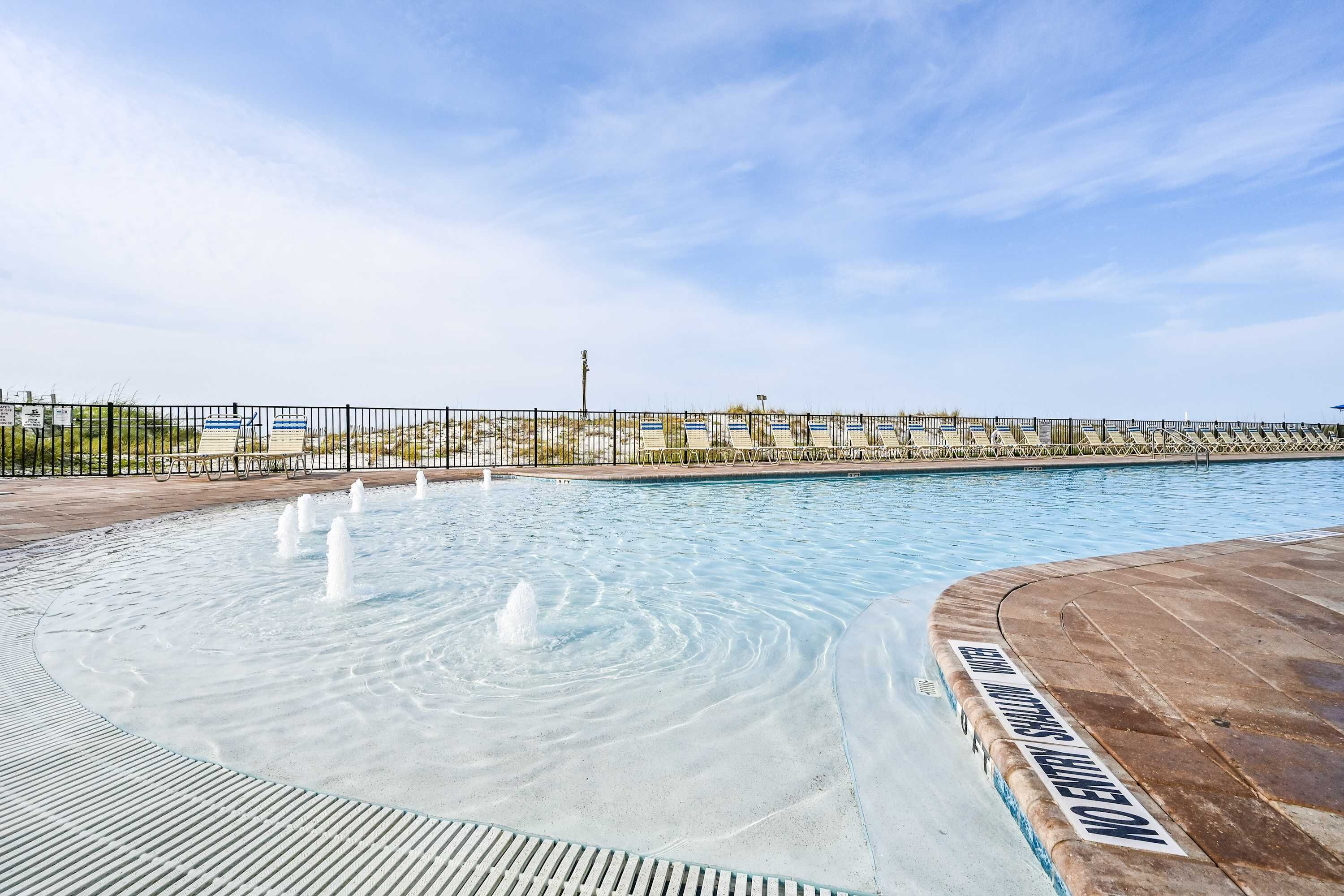 Ein Swimmingpool mit Springbrunnen, umgeben von einem Zaun und wolkenlosem Himmel.