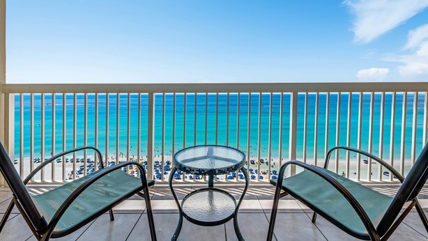 Een balkon met een glazen tafel en twee stoelen, met uitzicht op een strand met parasols en mensen.