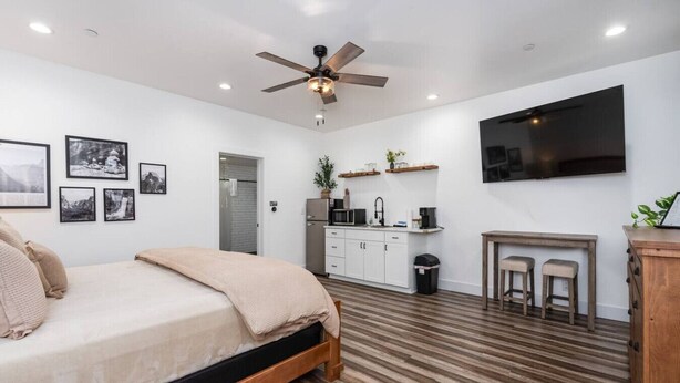 A modern bedroom with a wooden floor, a bed with a beige blanket, a ceiling fan, and a kitchen area with a TV and bar stools. - Exquisite Chic King Suite at Yosemite`s Doorstep (Oakhurst)