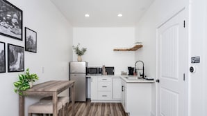 Une cuisine compacte avec des meubles blancs, un réfrigérateur, un four à micro-ondes et un évier. On y trouve une table en bois avec un banc et une plante en pot.
