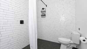 Une salle de bain avec des carreaux de céramique blancs rectangulaires, des toilettes blanches, une poubelle noire et un rideau de douche.
