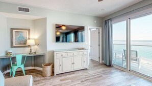 Een ruime kamer met een grote schuifglazen deur die uitkomt op een balkon, een houten vloer, een witte kast, een blauwgroene fauteuil en een ingelijst kunstwerk aan de muur.
