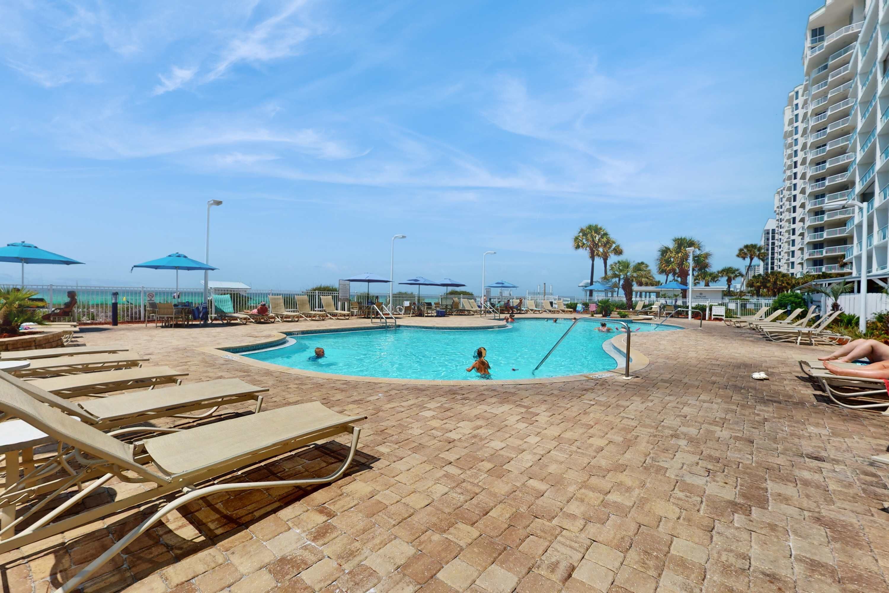 A pool area with lounge chairs and umbrellas, surrounded by a brick patio and a tall building.
