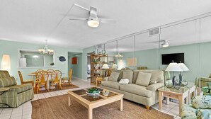 Ein Wohnzimmer mit Sofa, Couchtisch und Sesseln. Es gibt eine Essecke mit Tisch und Stühlen. Der Raum verfügt über einen Deckenventilator und einen an der Wand befestigten Fernseher.