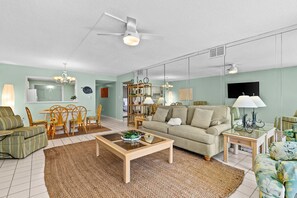 Un salon comprenant un canapé, une table basse et des fauteuils. On y trouve également un coin repas avec une table et des chaises. La pièce est équipée d’un ventilateur de plafond et d’un téléviseur fixé au mur.